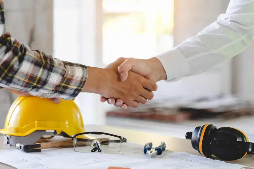 A contractor and property owner shaking hands beside project plans and safety equipment.