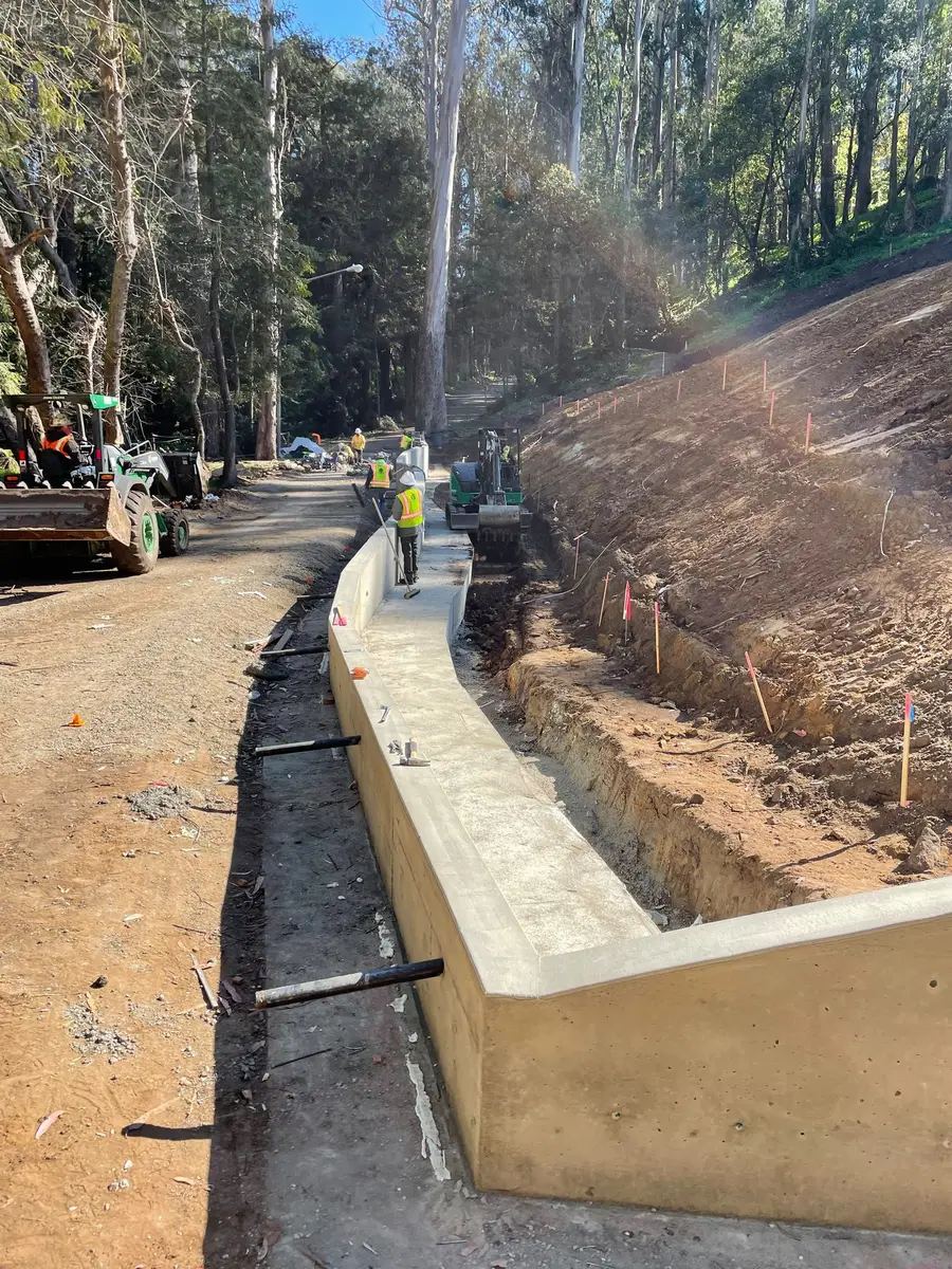 Poured concrete retaining wall following a hillside cut, with the crew and excavator working the line.